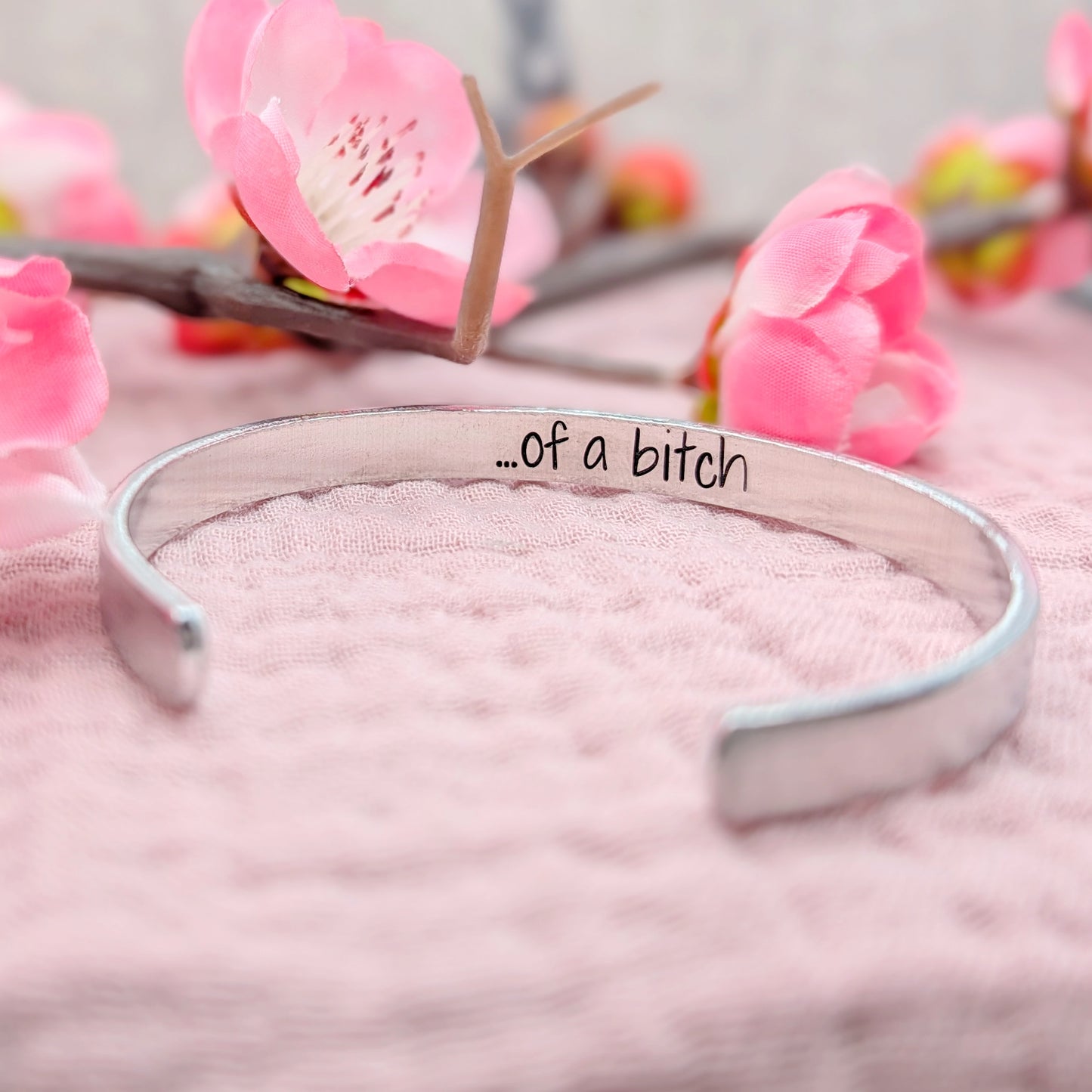 Silver bracelet with 'of a bitch' handstamped on the inside on a pink surface with flowers in the background