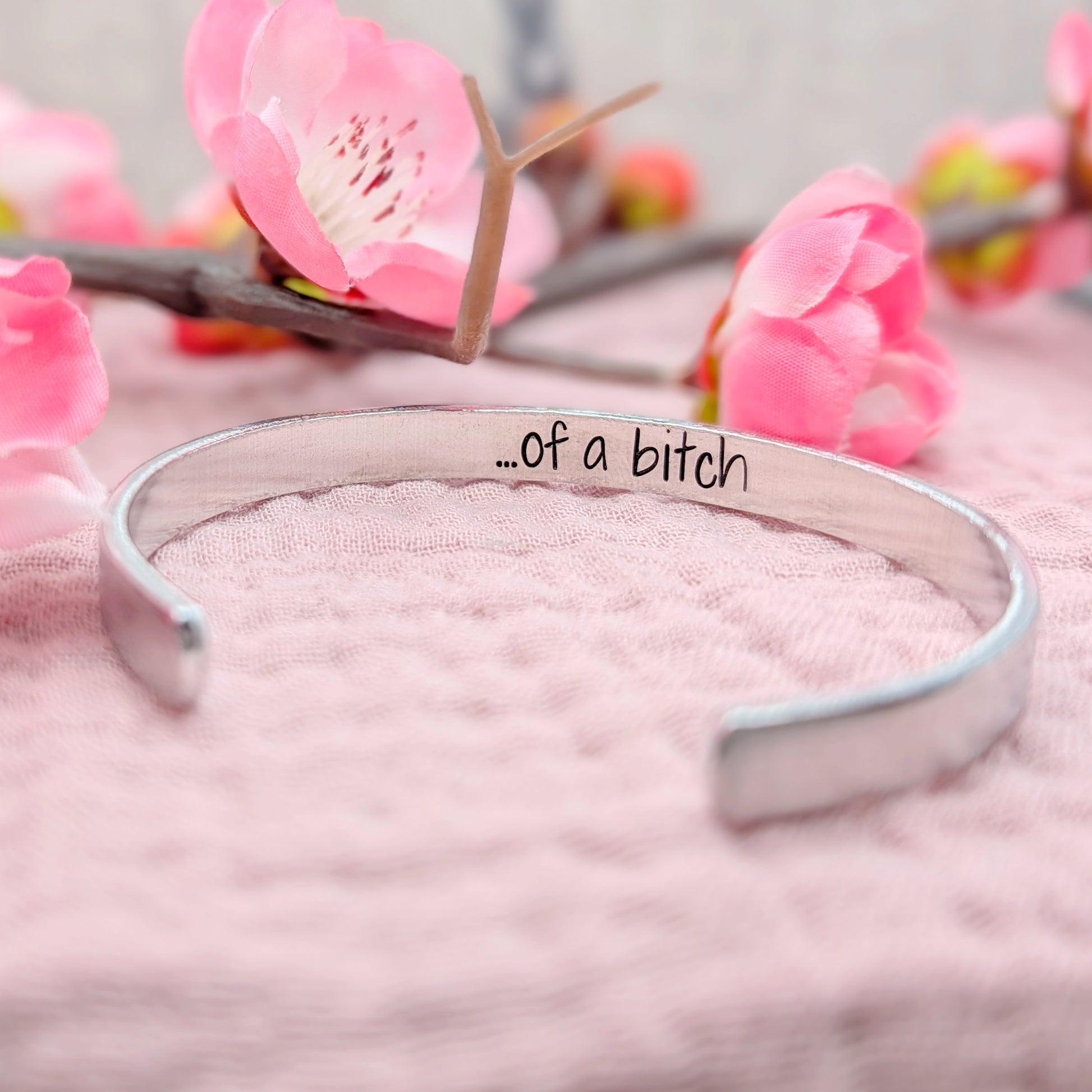 Silver bracelet with 'of a bitch' handstamped on the inside on a pink surface with flowers in the background